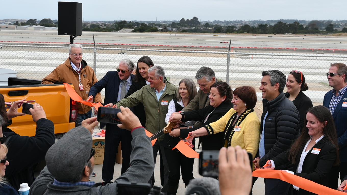 MRY Staff and Partners cut orange ribbon with Construction site and Airfield in background.